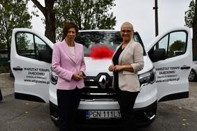 Pokaż zdjęcie: Dwie kobiety stoją przed mikrobusem w kolorze białym z otwartymi przednimi drzwiami. Na drzwiach widnieje napis: „Warsztat Terapii Zajęciowej”, adres placówki oraz adres jej strony internetowej. W tle są widoczne drzewa liściaste.