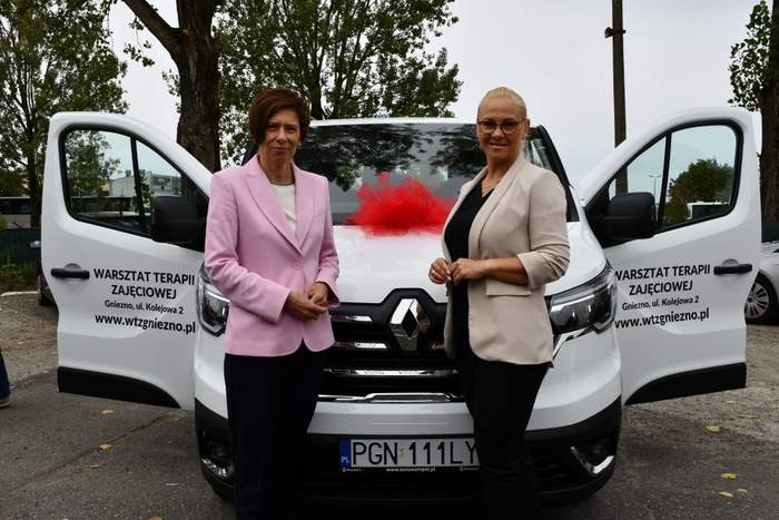 Pokaż zdjęcie: Dwie kobiety stoją przed mikrobusem w kolorze białym z otwartymi przednimi drzwiami. Na drzwiach widnieje napis: „Warsztat Terapii Zajęciowej”, adres placówki oraz adres jej strony internetowej. W tle są widoczne drzewa liściaste.