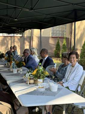 Pokaż zdjęcie: Grupa siedmiu osób siedzi pod zadaszonym namiotem przy stolikach ustawionych w jednym rzędzie. Na stolikach ustawione są napoje, cukierki oraz bukiety słoneczników.