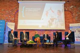 Pokaż zdjęcie: na zdjęciu w tle duży telebim z logo konferencji po środku siedzą zaproszeni goście i debatują na temat problemów osób z niepełnosprawnościami 