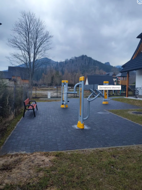 Pokaż zdjęcie: Trzy stacje siłowni plenerowej, po lewej stronie ławka, po prawej stronie budynek.