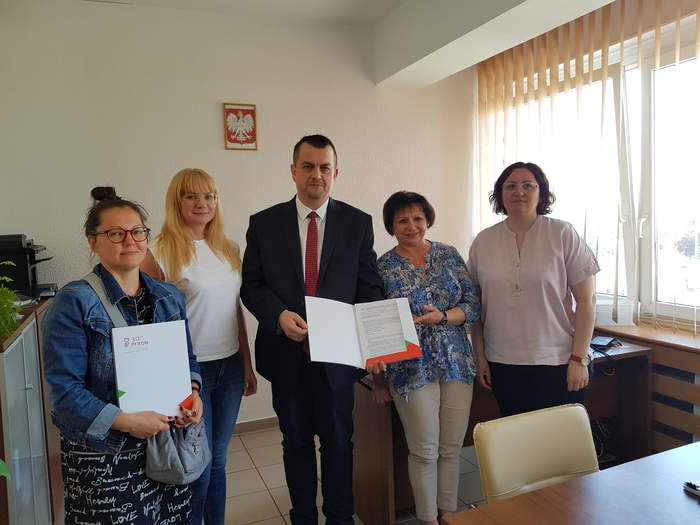 Pokaż zdjęcie: W Gabinecie Dyrektora stoją osoby podpisujące umowę i odpowiedzialne za jej realizację. Dyrektor Oddziału wspólnie z Prezes Stowarzyszenia trzymają umowę. Wszyscy stoją na tle ściany