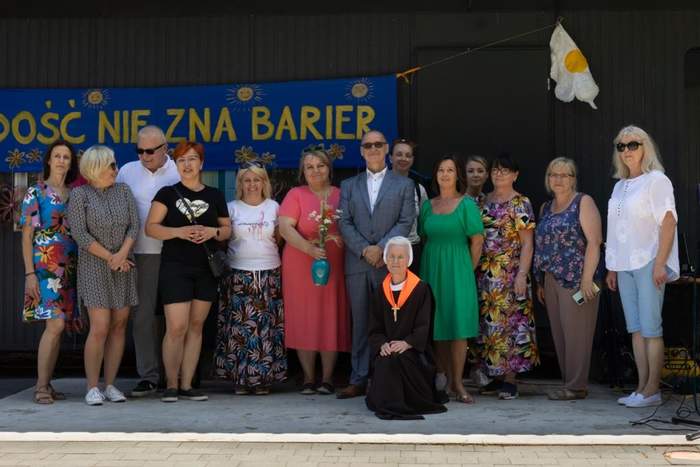 Pokaż zdjęcie: Czternaście osób pozuje do pamiątkowej fotografii, na tle napisu radość nie zna barier.