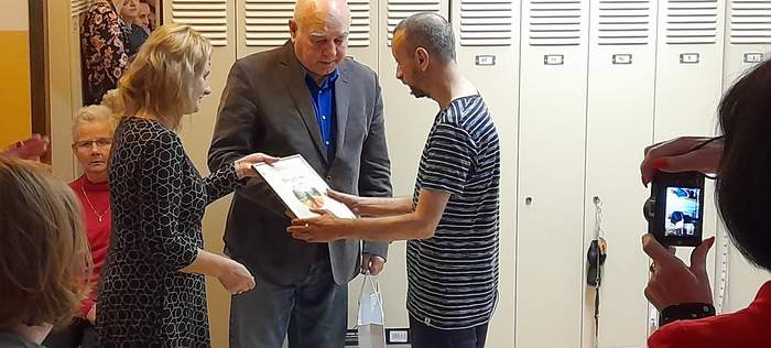 Pokaż zdjęcie: Na zdjęciu kobieta blondynka i starszy mężczyna wręczaja dyplom młodemu mężczyźnie ubranemu w koszulkę z krótkim rękawem. Zdjęcie zrobione w  sali.