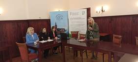 Pokaż zdjęcie: Kobieta z mikrofonem w ręku w trakcie przemówienia, a obok niej trzy inne kobiety siedzące przy laptopie