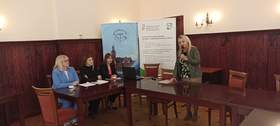 Kobieta z mikrofonem w ręku w trakcie przemówienia, a obok niej trzy inne kobiety siedzące przy laptopie