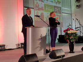 Pokaż zdjęcie: Mężczyzna w garniturze i krawacie przemawia przy mównicy, obok stoi kobieta w czarnej bluzce i granatowych spodniach, tłumacz języka migowego. Po prawej stronie duży ekran z kolorową grafiką. Przed kobietą stoi czarny wazon z białymi i czerwonym