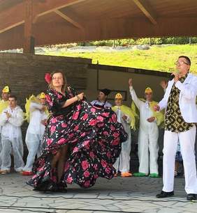 Pokaż zdjęcie: Występ Podopiecznych ze Stowarzyszenia Nasz Dom. Na scenie na pierwszym planie tancerka w hiszpańskiej sukni, obok mężczyzna z mikrofonem, za nimi grupa osób z niepełnosprawnościami w strojach kaczek 