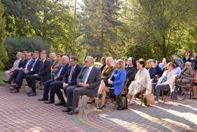 Pokaż zdjęcie: Na zewnątrz ośrodka, na krzesłach ustawionych w rzędach, siedzą zaproszeni goście, w pierwszym rzędzie mężczyźni w garniturach.