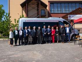 Grupa kilkunastu osób na tle busa, stoją w szeregu, za samochodem widać budynek