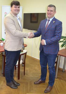 Pokaż zdjęcie: Dwóch mężczyzn podaje sobie dłoń. Są w gabinecie, za nimi stół, telewizor wiszący na ścianie i rollup PFRON