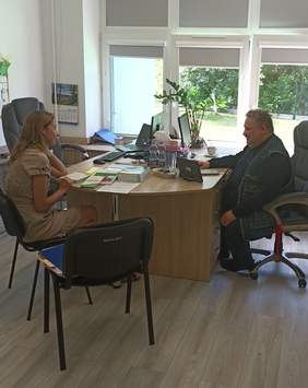 Pokaż zdjęcie: Mężczyzna i kobieta siedzą przy biurku, na którym znajduje się m.in. komputer, tablet, ulotki, poradniki, broszury. Uśmiechnięta kobieta z zaciekawieniem słucha mężczyzny