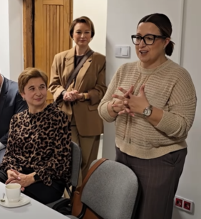 Pokaż zdjęcie: Przy stole stoi kobieta i składa życzenia z okazji otwarcia nowego budynku, pozostałe osoby siedzą zasłuchane.