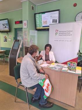 Pokaż zdjęcie: Przy biurku siedzi kobieta oraz mężczyzna, rozmawiają. Na stole leżą ulotki i foldery.