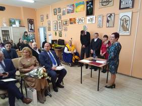 Pokaż zdjęcie: W sali domu opieki na krzesłach siedzą zaproszeni goście, w pierwszym rzędzie mężczyźni w garniturach oraz kobieta w beżowej sukience trzymająca kwiaty, na drugim planie widać osoby stojące przy ścianie