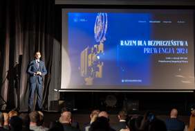 Pokaż zdjęcie: Mężczyzna w garniturze stoi na scenie przed dużym ekranem, na którym wyświetlony jest ten sam tekst: "RAZEM DLA BEZPIECZEŃSTWA - PREWENCJA 2024". Wygląda na to, że mężczyzna wygłasza przemówienie lub prowadzi wydarzenie. Sala jest wypełniona ludźmi siedzącymi na krzesłach, uważnie słuchającymi prelegenta.