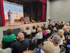 Widok na salę widowiskową, gdzie na scenie za jasną mównicą przemawia osoba w niebieskim stroju. Za nią, na dużym ekranie, wyświetla się napis 35 lat Towarzystwa Pomocy Potrzebującym im.. św. Alberta „NADZIEJA”. Scenę otaczają czerwone kurtyny, a obok stoi kamera na statywie. Na widowni w czarnych krzesłach siedzi elegancko ubrana, głównie starsza publiczność.