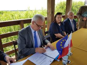 Pokaż zdjęcie: 5 osób zgromadzonych na tarasowej zabudowie, wokół ustawionego tam stołu z flagami Unii Europejskiej i Polski. W centralnej części następuje uroczyste podpisywanie dokumentów przez mężczyznę w garniturze, obserwowane przez zgromadzonych.