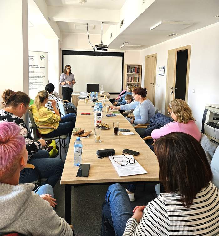 wzdłuż stołu konferencyjnego siedzą kobiety i słuchają prelegentki - przedstawicielki PFRON Oddziału Kujawsko-Pomorskiego, za którą stoi ekran, a po jej lewej stronie baner PFRON