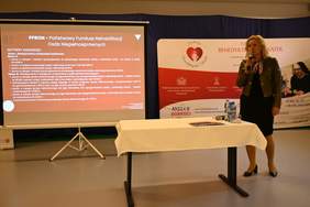 Pokaż zdjęcie: Przy mikrofonie stoi kobieta blondynka w zielonej ramonesce. Z tyłu slajd z prezentacji.