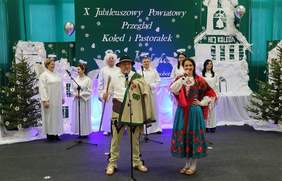 Pokaż zdjęcie: Na scenie ustawionej w świątecznej dekoracji występują artyści w strojach ludowych i anielskich. Z przodu stoją kobieta i mężczyzna śpiewający do mikrofonów, za nimi chór w białych strojach z anielskimi skrzydłami. W tle widoczny jest napis „X Jubileuszowy Powiatowy Przegląd Kolęd i Pastorałek Hej, Kolęda”, choinki oraz dekoracje nawiązujące do Bożego Narodzenia.