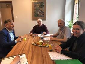 Pokaż zdjęcie: Przy stole siedzą trzej mężczyźni i kobieta uśmiechająca się do obiektywu. Na stole leżą dokumenty, pieczątki, woda mineralna szklanki i cukierki w talerzu.  W tle na białej ścianie wisi obraz.