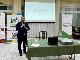 Pokaż zdjęcie: Mężczyzna w granatowym garniturze, krawacie i białej koszuli stoi trzymając w rękach mikrofon. Za nim ekran na którym wyświetlane jest logo PFRON.