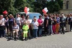 Pokaż zdjęcie: Grupa trzydziestu osób stoi w rzędzie przed białym busem, który udekorowany jest białymi i czerwonymi balonami. Z przodu znajdują się dwie kobiety, które na co dzień poruszają się na wózku. W tle widoczne są drzewa liściaste oraz fragment budynku.
