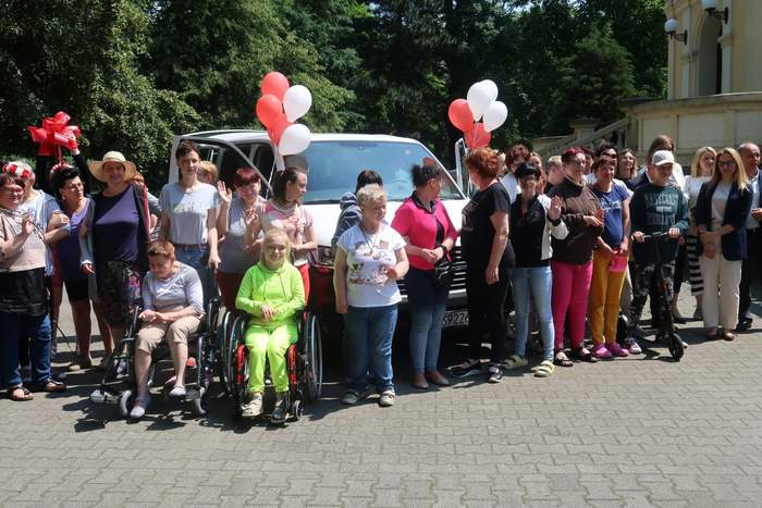 Pokaż zdjęcie: Grupa trzydziestu osób stoi w rzędzie przed białym busem, który udekorowany jest białymi i czerwonymi balonami. Z przodu znajdują się dwie kobiety, które na co dzień poruszają się na wózku. W tle widoczne są drzewa liściaste oraz fragment budynku.