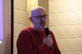 Pokaż zdjęcie: Zdjęcie mężczyzny w okularach z lekkim zarostem, ubranego w czerwony sweter. W ręku trzyma mikrofon