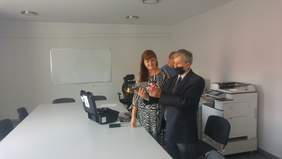 Pokaż zdjęcie: Pomieszczenie pomalowane na biało, na środku stoi duży biały stół z czarnymi krzesłami, przy stole stoi kobieta i mężczyzna, który trzyma w rękach drona i dokładnie go ogląda. Na stole leży czarna walizka.