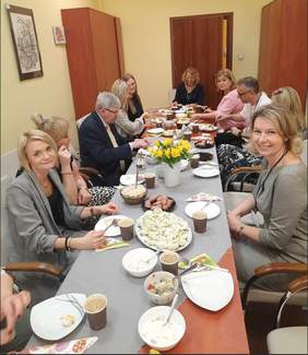 Pokaż zdjęcie: Wokół prostokątnego stołu siedzą ludzie. Stół zastawiony jest wielkanocnymi potrawami. Są na nim także świąteczne dekoracje. Ludzie jedzą, rozmawiają ze sobą i uśmiechają się. 