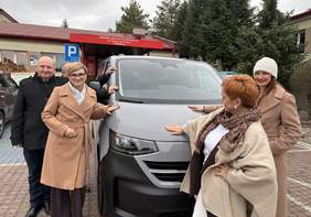 Pokaż zdjęcie: Grupa osób stoi przed nowym szarym mikrobusem zaparkowanym przed budynkiem Specjalnego Ośrodka Szkolno-Wychowawczego w Długoborzu. Uczestnicy uśmiechają się i symbolicznie dotykają pojazdu podczas jego przekazania placówce.