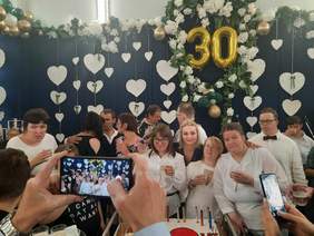 Pokaż zdjęcie: W pięknie przybranej sali stoją elegancko ubrani ludzie. Pozują do wspólnego zdjęcia. Przed nimi na stoliku leży ogromny tort.  