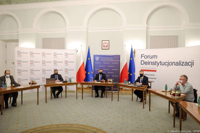 Pokaż zdjęcie: Przy stołach ustawionych w kształcie podkowy, znajdujących się w dużej, wysokiej sali siedzi pięciu mężczyzn. W środku Prezydent RP Andrzej Dude. W tle flagi polskie i unii europejskiej.