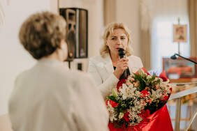 Pokaż zdjęcie: Zdjęcie dwóch kobiet, jedna trzyma w rękach kwiaty oraz mikrofon.