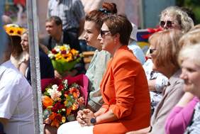 Pokaż zdjęcie: Uczestnicy wydarzenia siedzą na świeżym powietrzu; na pierwszym planie osoba w pomarańczowej marynarce trzyma bukiet kwiatów, wokół inni widzowie i dekoracje.