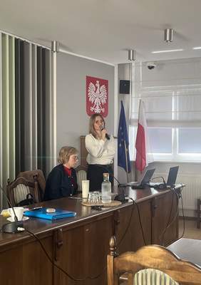 Pokaż zdjęcie: Kobieta przemawiająca do mikrofonu podczas spotkania Sieci Współpracy PFRON. Siedząca obok uczestniczka słucha wystąpienia. W tle widoczne godło oraz flagi Polski i Unii Europejskiej.