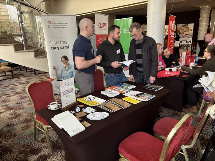 Pokaż zdjęcie: Dwóch mężczyzn w ciemnych koszulach stoi przy stoisku PFRON i rozmawia z mężczyzną w okularach i kurtce; na stole leżą broszury i materiały informacyjne.