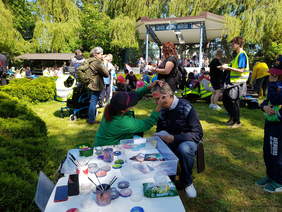 Pokaż zdjęcie: Przy stole z farbkami siedzi kobieta i mężczyzna. Kobieta w czapce z daszkiem maluje pędzlem twarz mężczyźnie. W tle scena i siedzący przed nią ludzie.