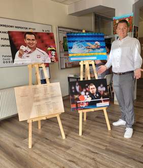 Pokaż zdjęcie: Po prawej stoi mężczyzna z rozłożonymi rękoma skierowanymi na cztery fotografie umieszczone na sztalugach. Na zdjęciu nr 1 mężczyzna w dłoni trzyma medal, nr 2 kobieta płynie w basenie, nr 3 lista zdobywców medali na olimpiadzie i Tokio, 
