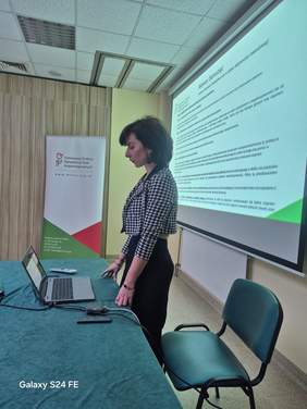 Pokaż zdjęcie: Prelegent kobieta stoi przy biurku, ręce oparte o blat. Kobieta ubrana jest w czarną spódnicę i marynarkę w pepitkę.