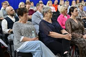 Pokaż zdjęcie: Dwie kobiety siedzą na widowni podczas Konferencja „Technologie asystujące wspierające wczesne wspomaganie rozwoju dziecka”. W tle widać licznie zgromadzonych uczestników wydarzenia.