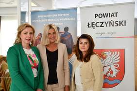 Pokaż zdjęcie: Do pamiątkowego zdjęcia pozują trzy kobiety na tle baneru z napisem powiat łęczyński