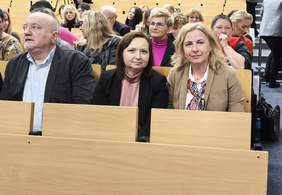 Pokaż zdjęcie: W auli, w ławkach siedzą uczestnicy konferencji. Zbliżenie na dwie kobiety, uśmiechające się do aparatu.
