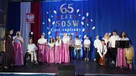 Na scenie występują dzieci z okazji 65-lecia Specjalnego Ośrodka Szkolno-Wychowawczego w Koluszkach. Dzieci śpiewają i grają na instrumentach. Mają na sobie eleganckie ubrania. 