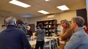 Pokaż zdjęcie: W pokoju przy stole stoją dwie kobiety i rozmawiają, po prawej stronie zdjęcia mężczyzna i kobieta zerkająca w stronę aparatu, po lewej stronie zdjęcia widać operatora kamery w słuchawkach, stojącego przy kamerze, znajdującej się na statywie