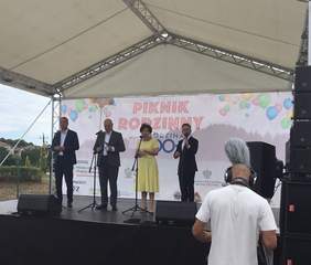 Pokaż zdjęcie: Trzech mężczyzn w garniturach wraz z jedną kobietą w żółtej sukience stoją na scenie. Każda z tych osób ma przed sobą oddzielny mikrofon na statywie. Na scenie ustawione zostały także kolumny głośnikowe oraz dwa odsłuchy. W tle, na białym ekranie wyświetlony został duży plakat promujący Piknik Rodzinny Rodzina 800 + w Dąbiu. Przed sceną widoczny jest mężczyzna w słuchawkach na głowie obsługujący kamerę. Po lewej stronie możemy dostrzec fragment ogrodzenia, zabudowania jednorodzinne oraz krzewy liściaste i iglaste.