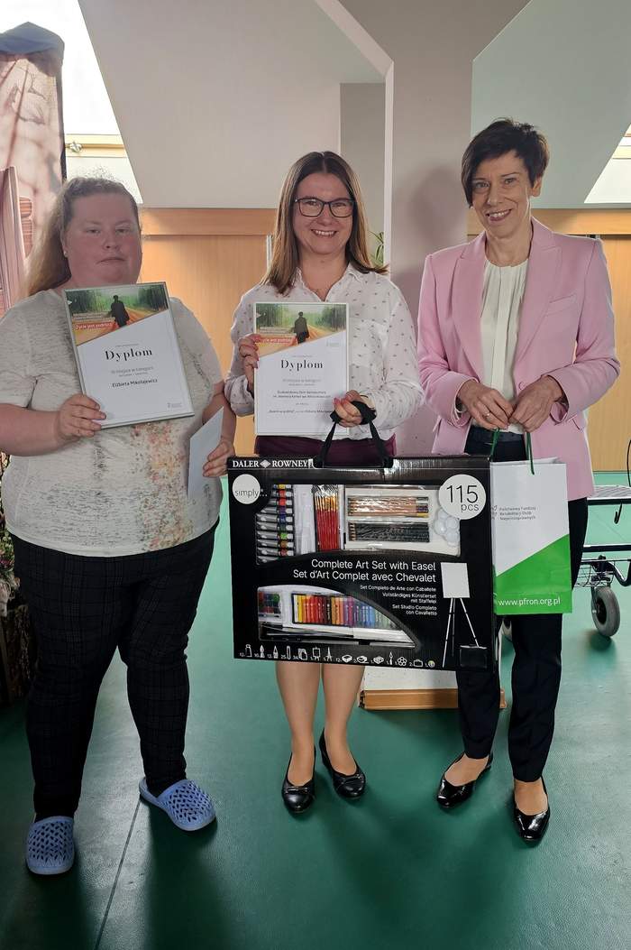 Pokaż zdjęcie: Trzy kobiety stoją w jednym rzędzie. Kobieta po lewej stronie trzyma w dłoniach dyplom oraz kartę podarunkową. Kobieta po środku trzyma dyplom dla placówki wraz z zestawem artystycznym dla laureatki. Kobieta po prawej trzyma z kolei torbę z logo PFRON.