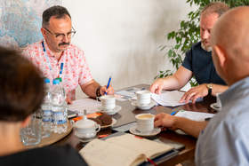 Kilka osób uczestniczy w spotkaniu przy stole konferencyjnym, analizując dokumenty i prowadząc rozmowę. Na stole znajdują się notatki, filiżanki z kawą oraz butelki wody.